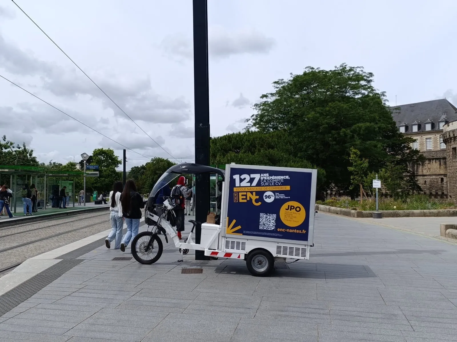 Triporteur publicitaire LCDG sur un parvis passant, près d’une station de tramway