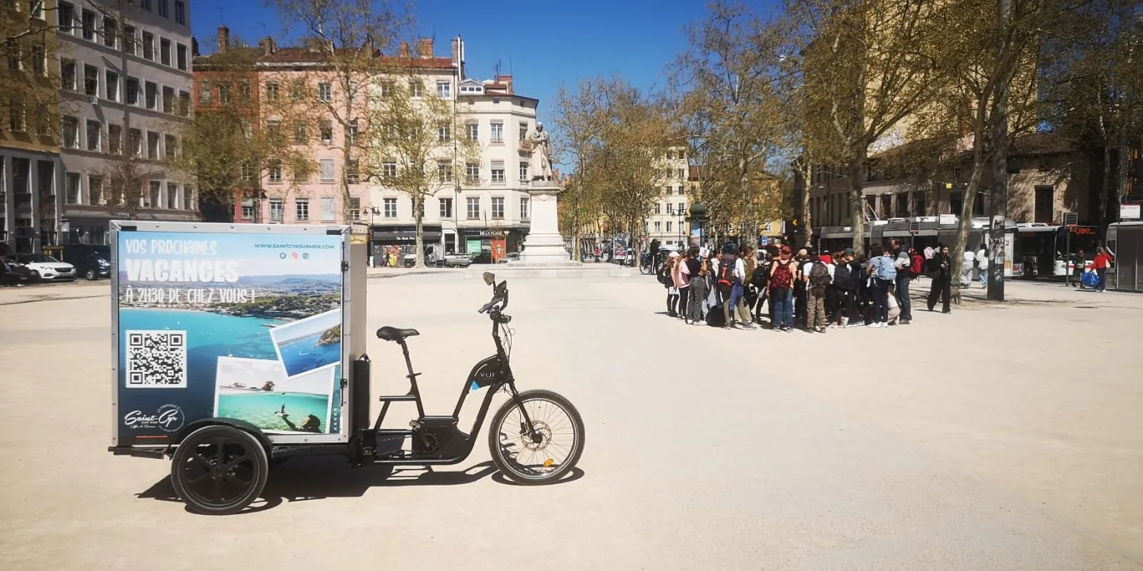 Vélo publicitaire LCDG sur une grande place piétonne, passants en arrière-plan