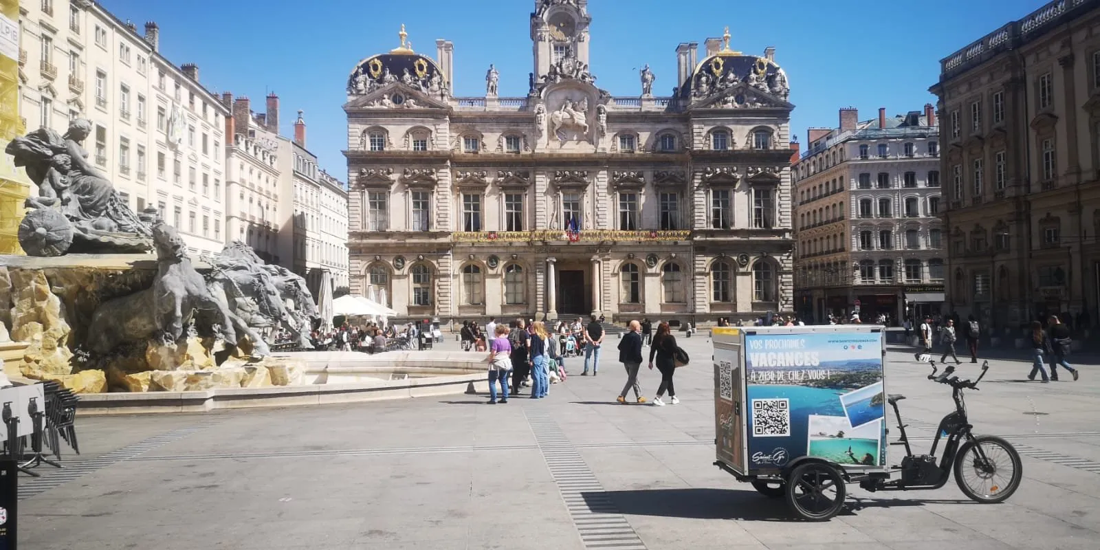 Vélo publicitaire LCDG sur une grande place ensoleillée en centre-ville